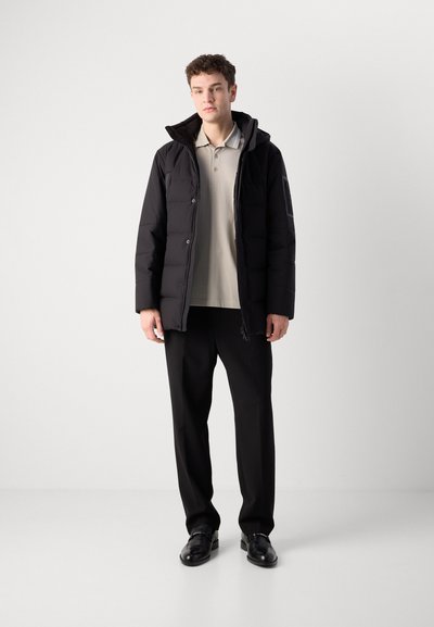 Doudoune noire avec capuche, boutons sur le devant et poches latérales, portée sur une chemise polo beige clair et un pantalon noir, accompagnée de chaussures noires.