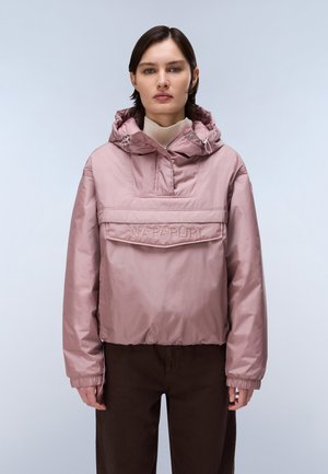 Femme portant une veste à capuche mauve clair avec une fermeture éclair à l’avant et une grande poche, associée à un pantalon marron foncé, debout devant un fond uni.