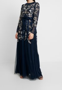 Marinblå golvlång klänning med långa genomskinliga Ärmar, prydd med silverfärgad blommönsterbroderi och paljetter, med ett knytband vid midjan.