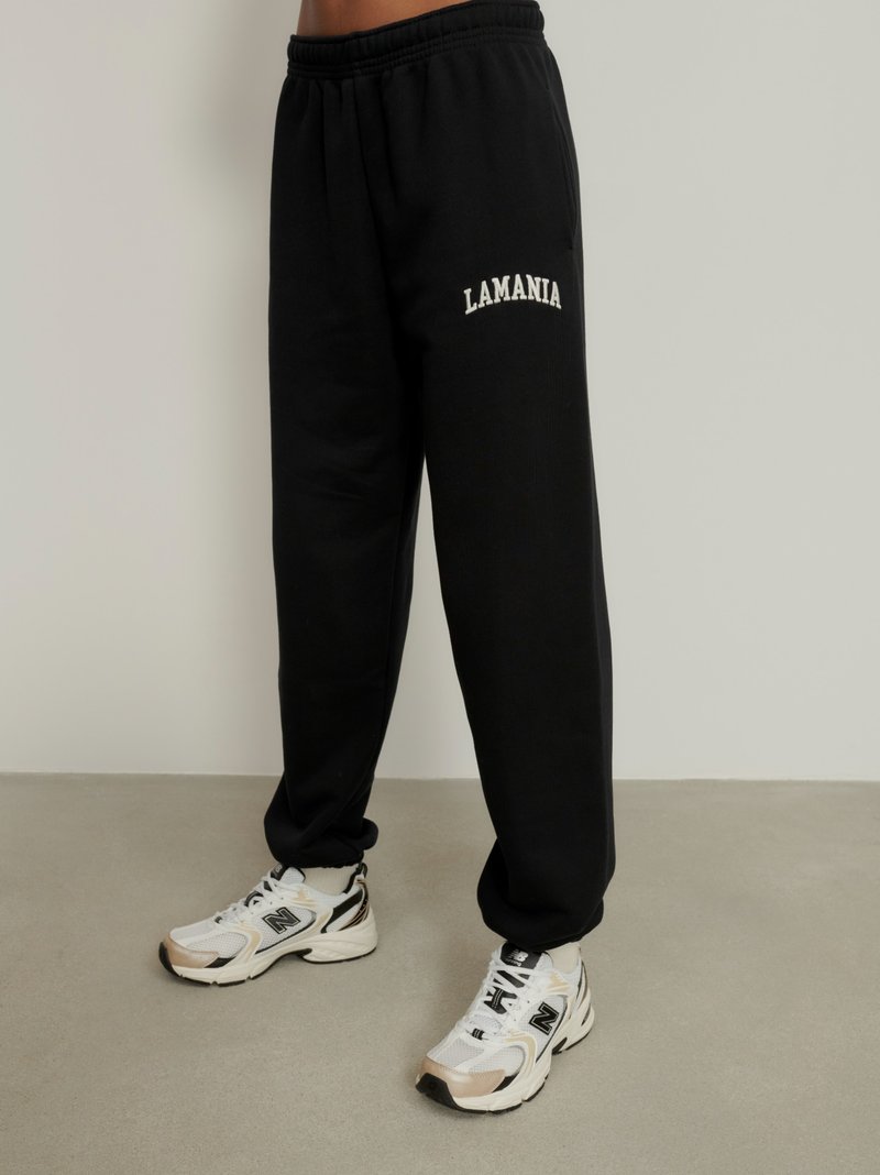 Čierne tepláky vyrobené z jemného materiálu s elastickým pásom. Na boku je vyšívané logo "LAMANIA" v bielej farbe. Skombinované s bielymi teniskami.