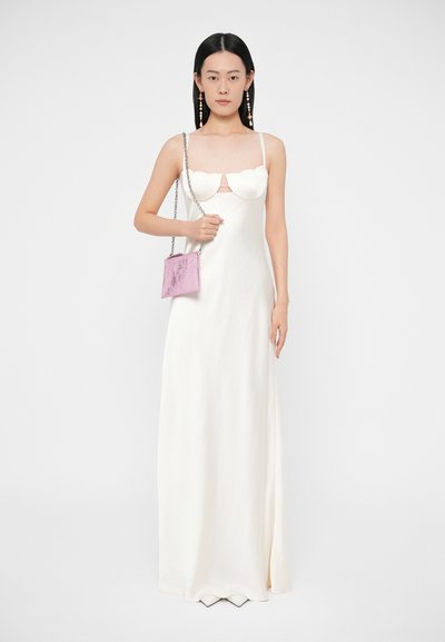 Sieviete baltā grīdas garuma kleitā ar viļņotu krūšu daļu, tur mazu rozā rokas somiņu ar sudraba ķēdes siksniņu, stāvot pret baltu fonu.