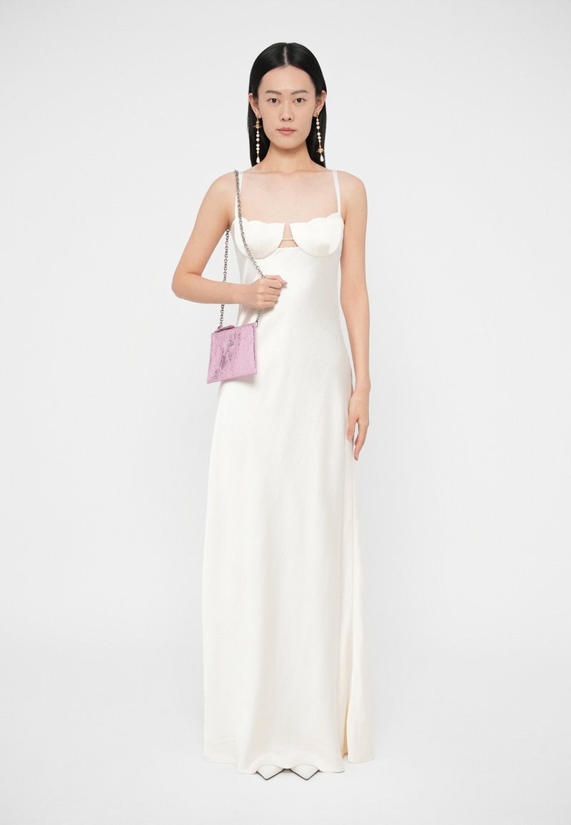 Mujer con un vestido blanco hasta el suelo con escote en forma de concha, sosteniendo un pequeño bolso rosa con cadena plateada, de pie contra un fondo blanco.