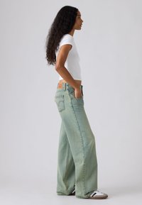 Γυναίκα με μακριά κατσαρά μαλλιά, φοράει λευκό εφαρμοστό μπλουζάκι, ανοιχτό μπλε τζιν παντελόνι με φαρδιά μπάτ και λευκά αθλητικά παπούτσια με μαύρες ρίγες, στέκεται προφίλ.