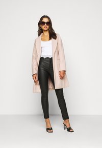 Lichtroze lange jas over een witte tanktop met kanttrim, gecombineerd met zwarte high-waisted leggings en zwarte open teen hakken. Grote zwarte zonnebril.