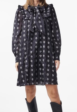 Schwarzes Langarmkleid mit weißem Blumenmuster, großer Schleife am Hals, geraffte Schulterdetails, kombiniert mit schwarzen knielangen Stiefeln.