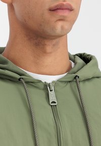 Lahka zelena zip-up jakna z mrežasto podloženim kapuco, ki ima izrazit zadrgo in naglavne akcentne vrvice. Gladka tekstura in sodoben kroj.