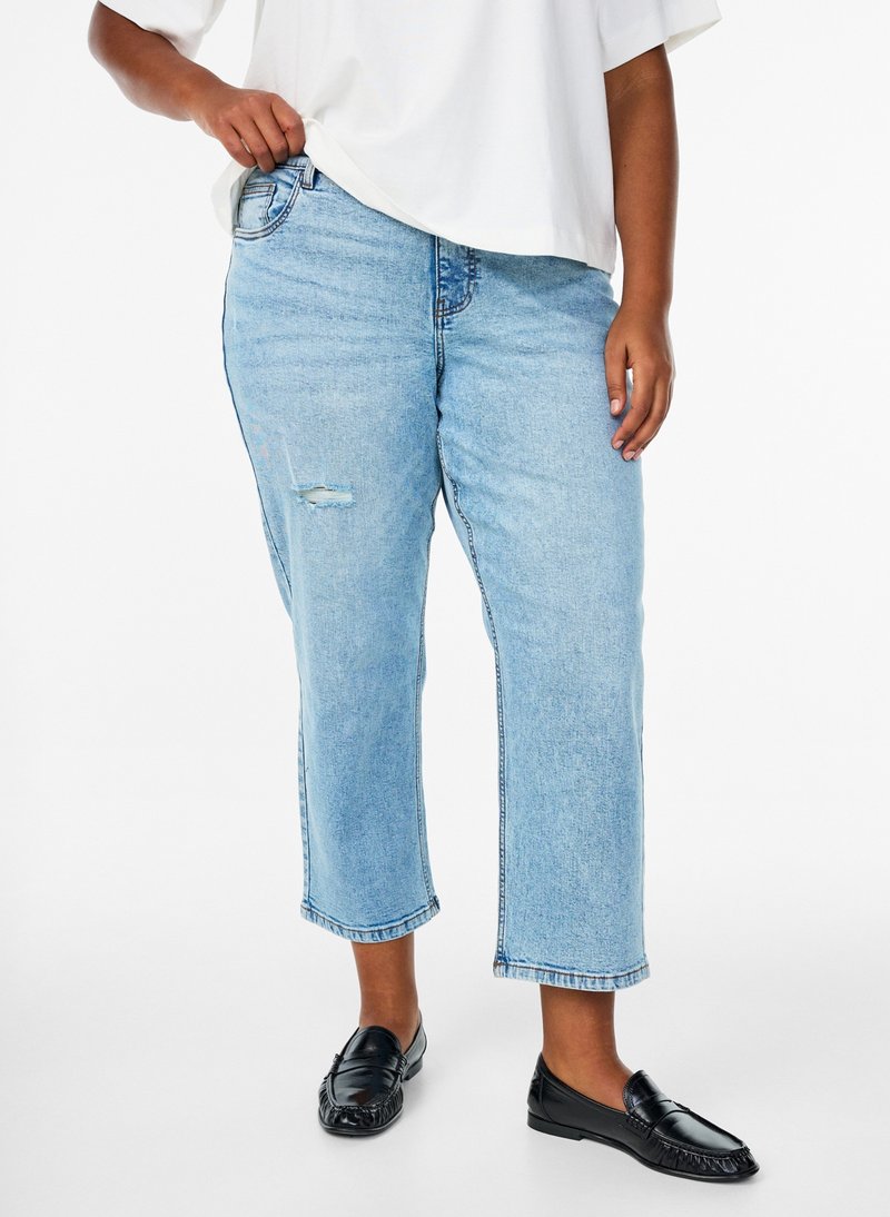 Person wearing light blue cropped jeans with a small rip on the thigh, a white top, and black slip-on loafers, standing against a white background.