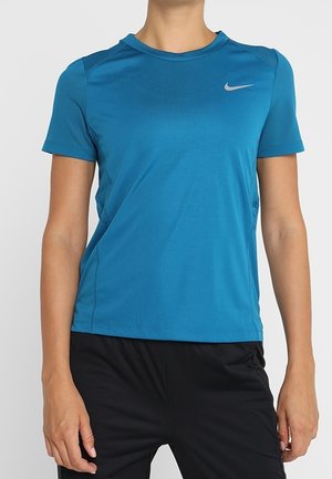 Osoba w niebieskiej sportowej koszulce Nike z krótkim rękawem i czarnych spodniach, pokazana od szyi do bioder na jasnym tle.