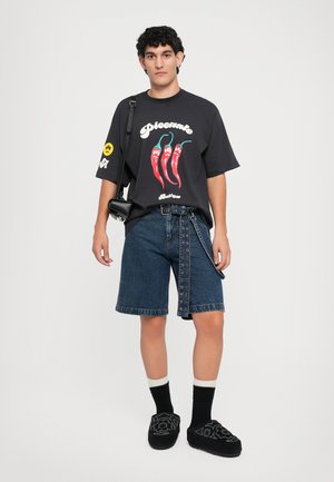 T-shirt nera oversize con grafica di peperoncino, abbinata a pantaloni shorts di denim blu con bottoni laterali e orli arrotolati. Scarpe nere slip-on.