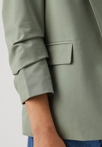 Světle zelený blazer se strukturovaným designem, hladkou texturou, detailem ohnutého rukávu a malou boční kapsou.