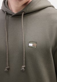 Gros plan sur une personne portant un sweat à capuche vert olive avec un patch logo "Tommy Jeans" sur la poitrine et des cordons suspendus à la capuche.