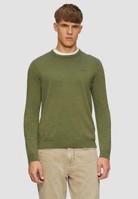 Grüner gestrickter Pullover mit Rundhalsausschnitt, gerippten Bündchen und Saum. Mit einem kleinen Logo auf der Brust. Kombiniert mit hellbeigen Hosen.