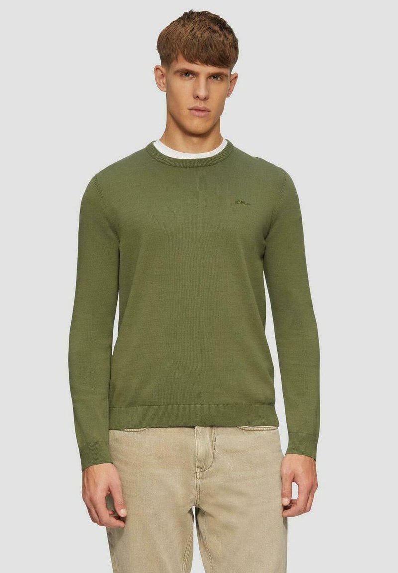 Grüner gestrickter Pullover mit Rundhalsausschnitt, gerippten Bündchen und Saum. Mit einem kleinen Logo auf der Brust. Kombiniert mit hellbeigen Hosen.