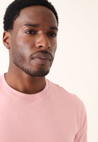 Lyserød bomulds-T-shirt med rund halsudskæring, med en blød tekstur og minimalistisk design. Nærbillede af ansigt, der viser huddetaljer og hår.