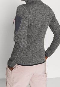 Kurtka polarowa w szarym kolorze o teksturowanym wzorze jodełki, z wysokim kołnierzem i niebieską łatą na ramieniu. Posiada zapinaną na zamek kieszeń na ramieniu.