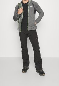 Giacca grigia con zip, pannelli neri e zip verde, abbinata a pantaloni neri e sneakers nere. Zaino appoggiato su una spalla.