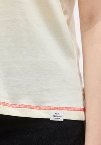 Λευκό πουκάμισο με κόκκινες ραφές στο τελείωμα και ετικέτα Mads Nørgaard Copenhagen, φορεμένο πάνω από σκούρο παντελόνι σε σώμα ατόμου.