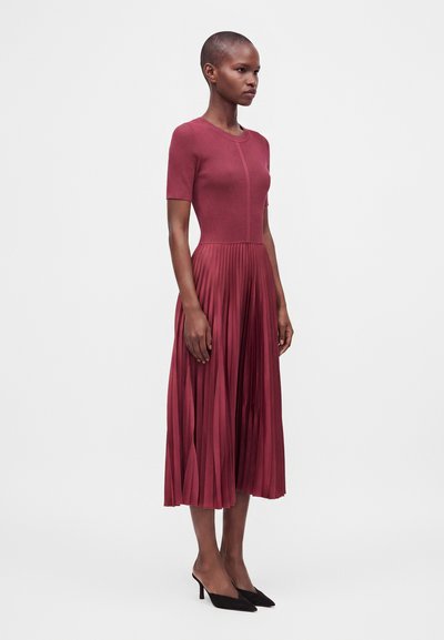 Femme portant une robe courte à manches courtes côtelée avec une jupe midi plissée couleur rose profond et des talons noirs pointus, debout de profil.
