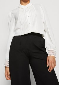 Blouse blanche avec un devant en dentelle texturée et des détails de boutons. Manches longues avec des accents de dentelle aux poignets, associée à un pantalon noir.