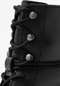 Närbild på svart texturerad läderstövel med svarta snören som löper genom silverfärgade metallkrokar.