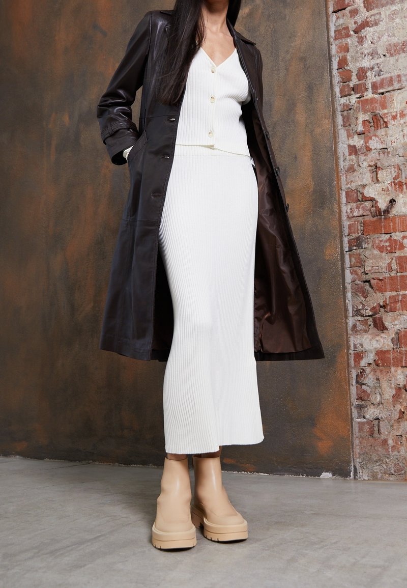 Svart lång kappa över en vit ribbad klänning och beige plattform ankle boots, som betonar en slank silhuett och strukturerade material.