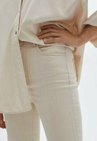 Jean en denim beige à coupe slim, avec coutures claires et un design classique à cinq poches. Une chemise légère et ample est partiellement rentrée.