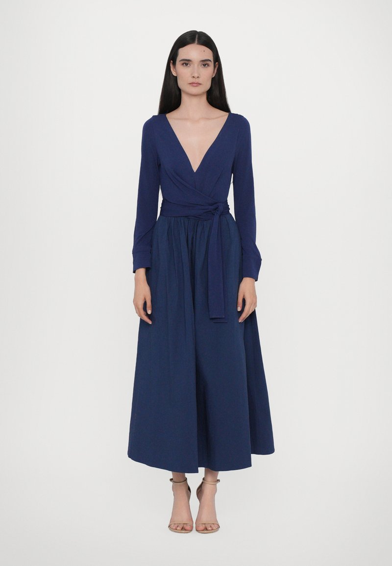 Navy blauwe wikkeljurk met lange mouwen, een V-hals en een striktaille. Vloeiende, volle rok met een gladde stoftextuur.