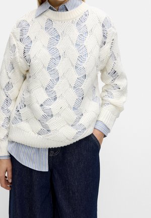 Personne portant un pull blanc en maille ajourée par-dessus une chemise à rayures bleues et blanches boutonnée, ainsi qu'un jean bleu foncé à jambes larges.