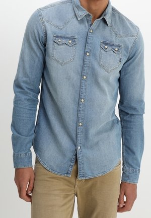 Mand iført en lyseblå denim skjorte med to brystlommer foran og beige bukser mod en ensfarvet baggrund.