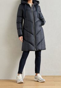 Abrigo acolchado de marina con acolchado en forma de chevron, cierre frontal y capucha adjunta. Combinado con jeans oscuros y zapatillas blancas con acentos plateados.