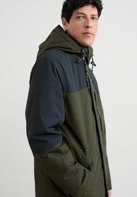 Bărbat îmbrăcat într-o jachetă cu glugă în două tonuri, verde și bleumarin, cu șireturi, privind peste umăr, cu mâna în buzunar, pe un fundal simplu.