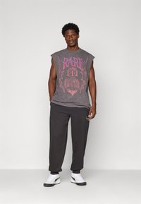 Ung mand med kort, krøllet hår iført en ærmeløs grå grafisk t-shirt, sorte joggers og hvide sneakers, der står foran en ensfarvet baggrund.