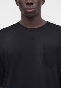Κοντινό ενός ατόμου που φοράει μαύρο t-shirt με στρογγυλή λαιμόκοψη και μικρό μαύρο κεντημένο λογότυπο πάνω από την αριστερή τσέπη στο στήθος.