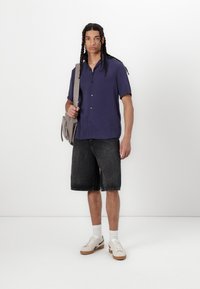 Kortærmet marineblå skjorte, sorte denimshorts og beige sneakers med brune såler. Model bærer en grå crossover-taske. Tekstureret stof.