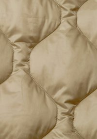 Pikowana kołdra w beżowym satynowym wykończeniu; charakteryzuje się diamentowym wzorem z gładką fakturą i misternymi detalami przeszyć.