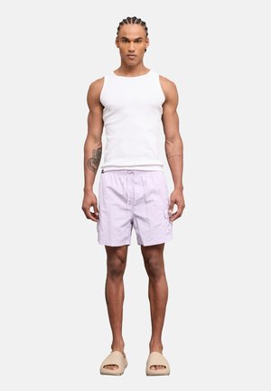 Muskelöser Mann mit geflochtenen Haaren, trägt ein weißes ärmelloses Shirt, hellviolette Shorts und beige Slidesandalen, steht frontal.