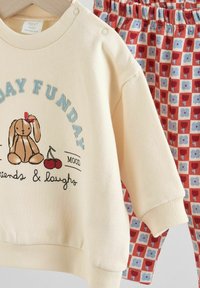 Cremefarbene Sweatshirt mit Hasenmotiv und Text, kombiniert mit rot gemusterten Hosen, die blaue und rote Quadrate zeigen. Aus weicher Baumwolle.