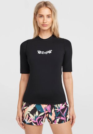 Frau trägt ein schwarzes kurzärmeliges Oberteil mit weißem Blumenmuster und "O'Neill"-Logo sowie bunte tropische Shorts, steht vor einem schlichten Hintergrund.