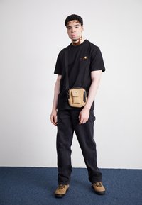 Black t-shirt with a small red and yellow logo, paired with dark jeans and tan footwear. A tan crossbody bag is worn across the body.