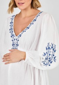 Weißes, strukturiertes Kleid aus Stoff mit V-Ausschnitt, verziert mit blauer Blumenstickerei entlang des Kragens und der Ärmel. Lange, gepuffte Ärmel mit elastischen Bündchen.