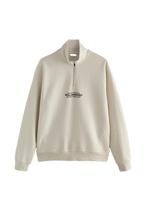 Beeži tooniga dressipluus poolest lukuga, kõrge krae, soonikkoes mansetid ja alumine äär, mille keskosas on musta värvi kiri: "STUDIO ORIGINAL WILLIAMSBURG."