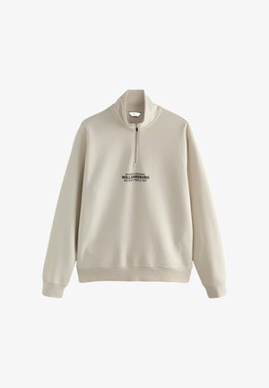 Beeži tooniga dressipluus poolest lukuga, kõrge krae, soonikkoes mansetid ja alumine äär, mille keskosas on musta värvi kiri: "STUDIO ORIGINAL WILLIAMSBURG."