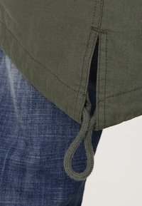 Detailní záběr na zelený lem tkaniny se bočním rozparkem a smyčkovou šňůrkou přes modré džíny.