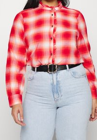 Kobieta ubrana w czerwoną i białą koszulę w kratkę zapinaną na guziki, włożoną w jasnoniebieskie spodnie z wysokim stanem, z czarnym paskiem.