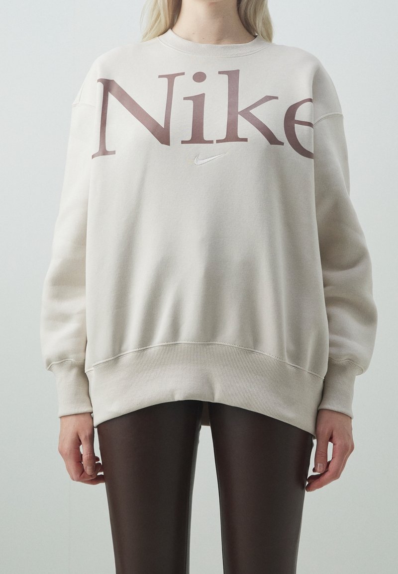 Světlá béžová mikina s velkým hnědým logem "Nike", kulatým výstřihem, spadlými rameny a žebrovanými manžetami a lemem, doplněná hnědými legínami.