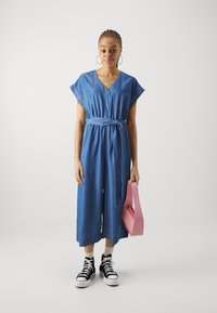 Une combinaison en denim avec un col en V, des manches courtes et une ceinture nouée à la taille ; associée à des baskets montantes noires et à un sac à main rose.
