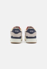 Sneakers met een lichtbeige bovenzijde, marineblauwe accenten en een textured zool. De achterkant toont het nummer "1933" in het wit.