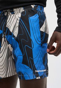 Κοντινό πλάνο ενός ατόμου που φορά μπλε, μαύρα και μπεζ σορτς με αφηρημένο μοτίβο, με το λογότυπο "Peak Performance", μαύρο κορδόνι και ελαστική μέση.