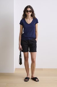 Jūras zila V-veida kakla īsām piedurknēm augšdaļa, kombinēta ar melniem šortiem. Modelis tur melnu roku somu un valkā melnas sandales ar mezgla dizainu.