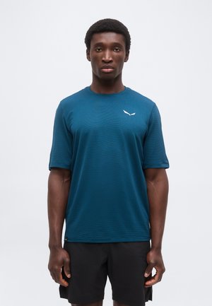 Noormees seisab ettepoole vaadates, seljas merevaiguroheline lühikeste varrukatega sportlik särk ja mustad lühikesed püksid tavalise valge tausta ees.